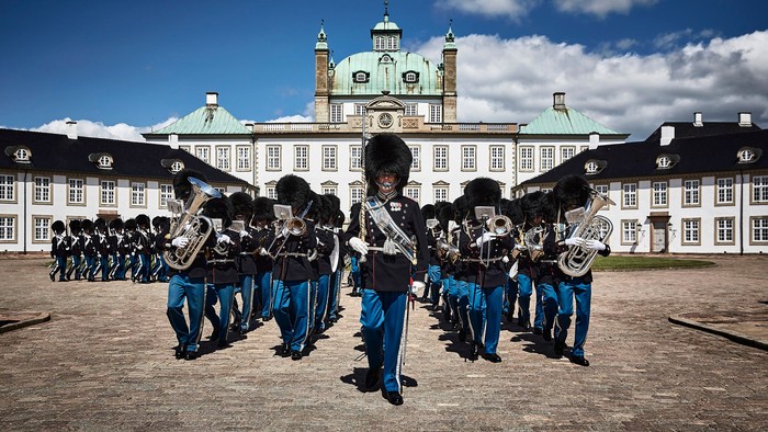 Uniformerede orkester medlemmer marcherer i formation med messinginstrumenter foran et elegant historisk palads.