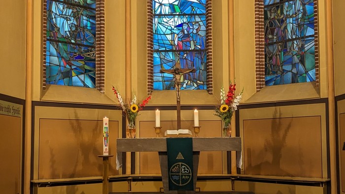 Apsis der Magdalenenkirche mit drei gestaltenten Fenstern und Altar mit Kreuz, Kerzen, Blumenschmuck und grünem Parament.