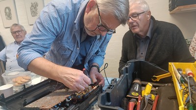 To mænd samarbejder tæt om at reparere en elektronisk enhed med værktøj og komponenter spredt rundt omkring.