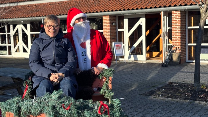 En person i julemandskostume sidder ved siden af en anden person på en festlig juleslæde udendørs.