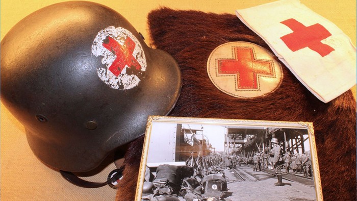 Historisk militærhjelm med medicinsk emblem og vintage slagmarksfotografi på pelsbaggrund.