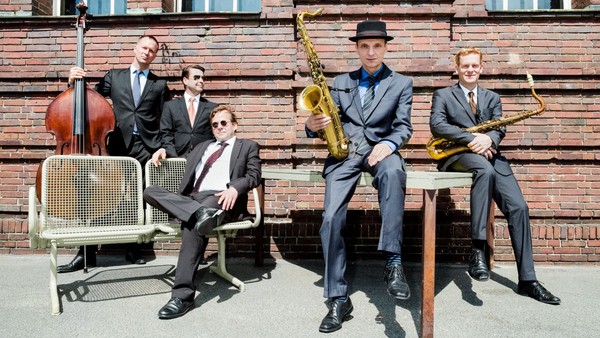 Ein Jazzquartett in formeller Kleidung posiert mit Instrumenten vor einer Ziegelsteinwand.