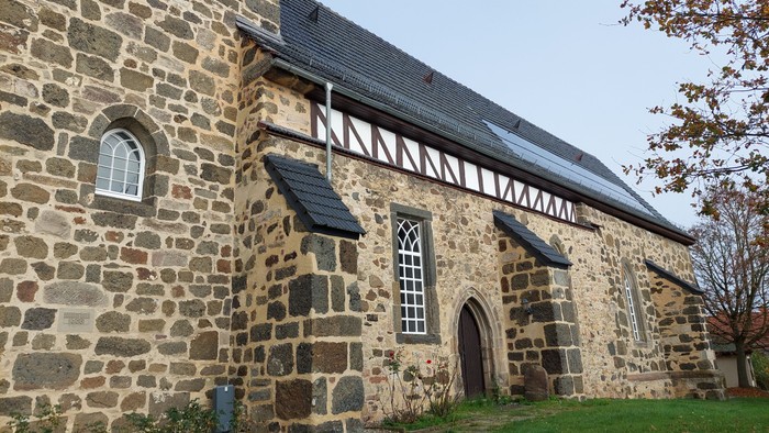 Steinkirche mit Bogenfenstern und -tür, umgeben von Gras und Bäumen.