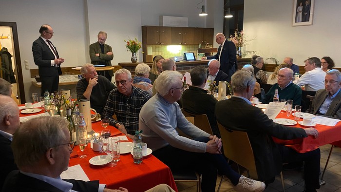 Gruppe af ældre mennesker samlet til en formel indendørs begivenhed med borde dækket til spisning og taler.