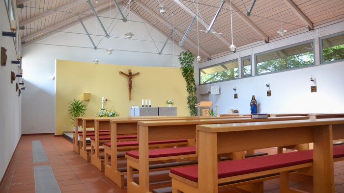 Moderner Kapelleninnenraum mit hölzernen Kirchenbänken, Altar und religiösen Dekorationen unter einer hohen, verglasten Decke.