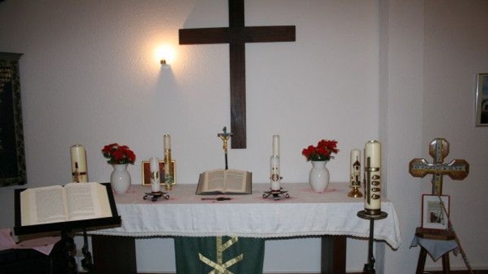 Eine ruhige Altargestaltung mit Kerzen, Büchern, Blumen und einem Kreuz auf einem bedeckten Tisch.