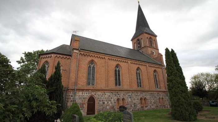 Historische Backsteinkirche mit hohem Turm und Bogenfenstern in einem ruhigen, landschaftlich gestalteten Friedhof.