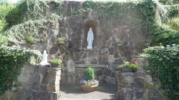 Steinheiligtum mit Statue der Jungfrau Maria, umgeben von üppigem Grün und farbenfrohen Blumentöpfen.