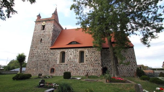 Historische Steinkirche mit rotem Ziegeldach und Uhrturm, umgeben von einem ruhigen Friedhof.