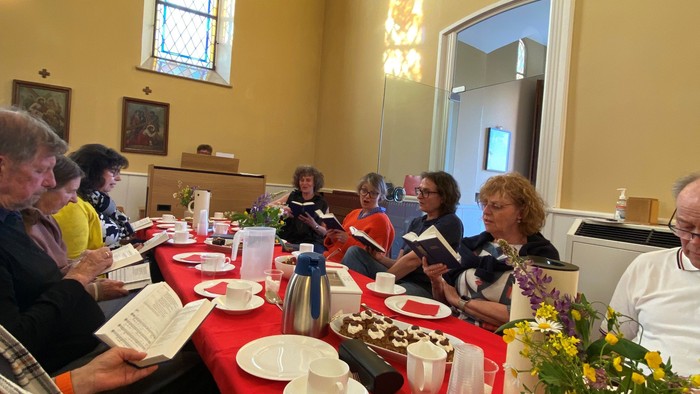 En gruppe af mennesker sidder ved et bord med mad og drikkevarer i et rum med et kors på væggen.