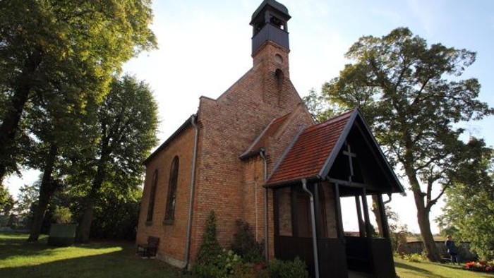 Idyllische Backsteinkirche mit Glockenturm, eingebettet zwischen grünen Bäumen und einer offenen Rasenfläche.