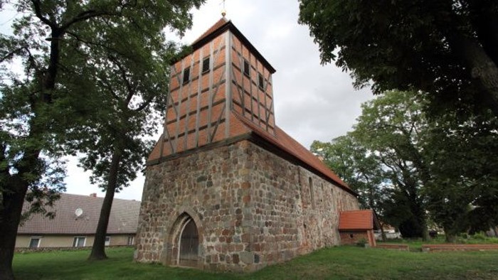 Historische Steinkirche mit Fachwerk-Glockenturm, eingebettet zwischen üppigen grünen Bäumen.