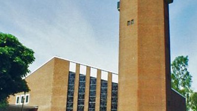 Das Bild zeigt einen hohen Ziegelbau mit einem Turm und umgeben von Grün.