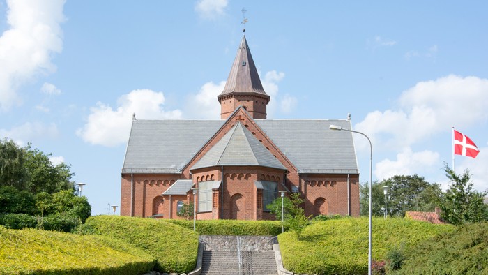 En rød kirke med spiret og en dansk flagvåbens flag på en klar dag