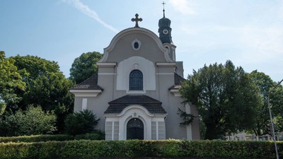 Kleine Kirche mit Kuppel und Kreuz auf dem Dach, umgeben von Bäumen und Büschen