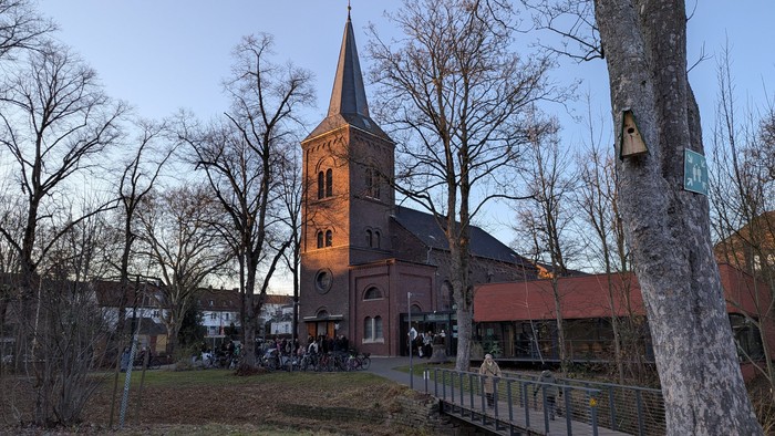 Eine historische Kirche mit hohem Turm, umgeben von kahlen Bäumen und einer kleinen Gruppe von Menschen in einem Außenbereich.