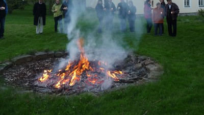 Die Gruppe versammelt sich um ein großes Lagerfeuer im Freien auf einer grasbewachsenen Fläche in der Nähe eines rustikalen Gebäudes.