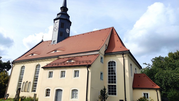 Ein weißes Kirchengebäude mit rotem Dach und hohem Turm, umgeben von Bäumen und einem Friedhof.