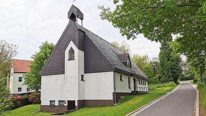 Kleine weiße Kirche mit schwarzem Dach und Kreuz, umgeben von Bäumen und einem Weg.