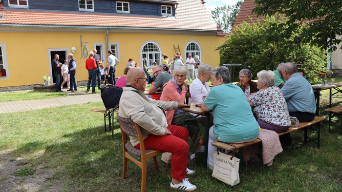 Senioren sitzen im Garten eines gelben Hauses auf Bänken und Stühlen und unterhalten sich.