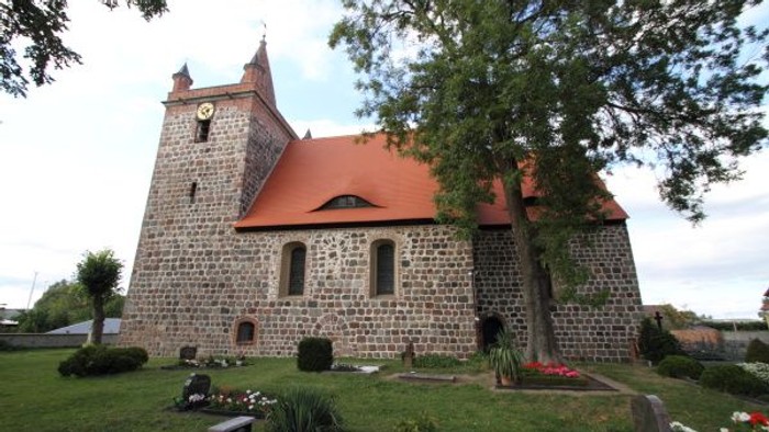 Historische Steinkirche mit rotem Ziegeldach und Uhrturm, umgeben von einem ruhigen Friedhof.
