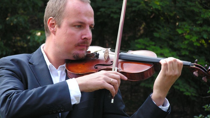 Ein Mann in formeller Kleidung spielt draußen in üppigem Grün Geige.