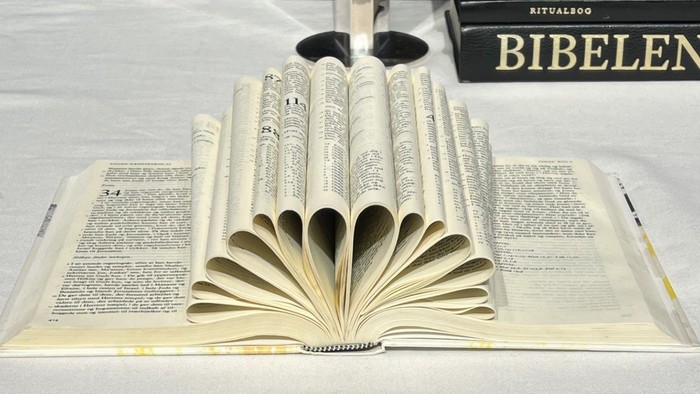 Bibel ligger åbnet på et alterbord - en del af siderne er foldet indad og danner en vifte. En anden bibel, en ritualbog og andre bøger i baggrunden