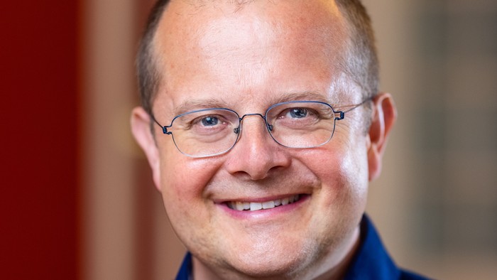 Mand med briller, der smiler, iført en blå knap-op skjorte indendørs.