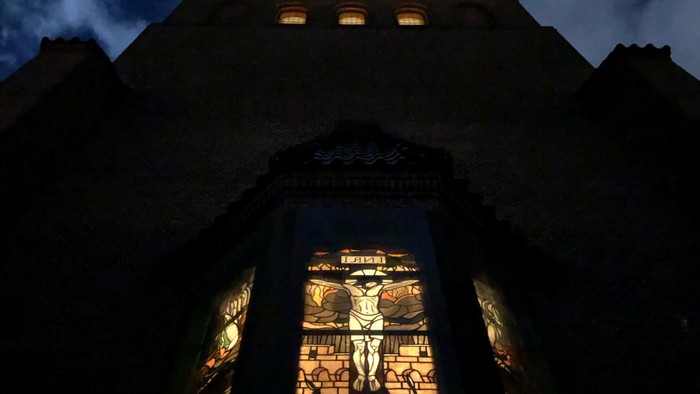 En stor, oplyst portbue om natten med et farvet glasvindue, der forestiller en korsfæstelsesscene.