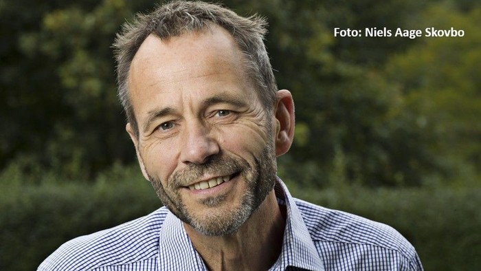 Mand med smil, kort gråt hår og skæg, iført en stribet knap-op skjorte udendørs.