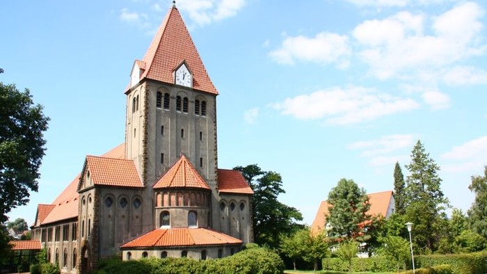 Das Bild zeigt eine große Kirche mit hohem Turm und rotem Dach, umgeben von grünem Rasen und Bäumen unter einem klaren blauen Himmel.