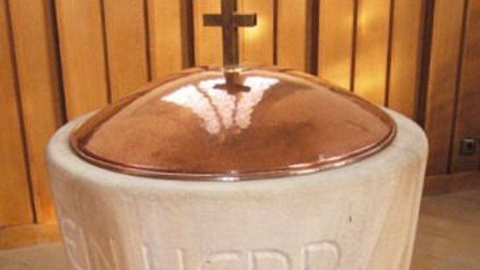 Der Taufstein mit Kreuz und lateinischer Inschrift in einer Kirche.