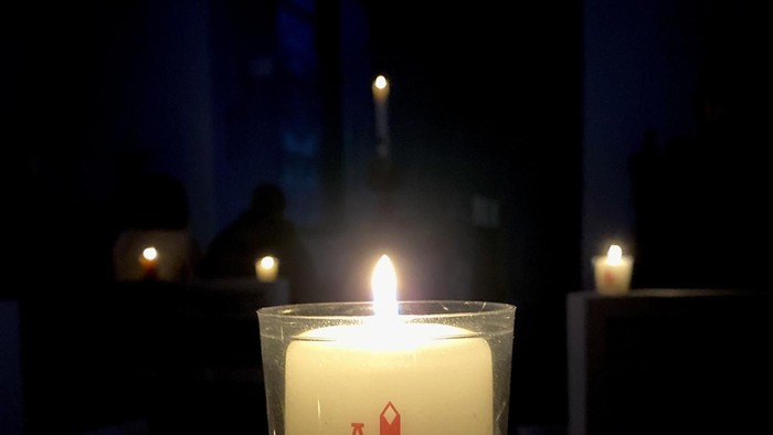 Eine brennende Kerze in einem Glasbehälter mit einem roten Kreuz und Omega-Symbol, die sanft in gedämpfter Umgebung leuchtet.