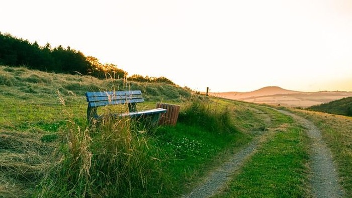 Eine einsame Bank steht auf einem grasbewachsenen Hügel mit Blick auf eine weite Landschaft bei Sonnenuntergang.