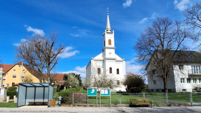 Ein weißer Kirchturm mit spitzem Dach steht neben einem gelben und einem grauen Gebäude unter einem klaren blauen Himmel.