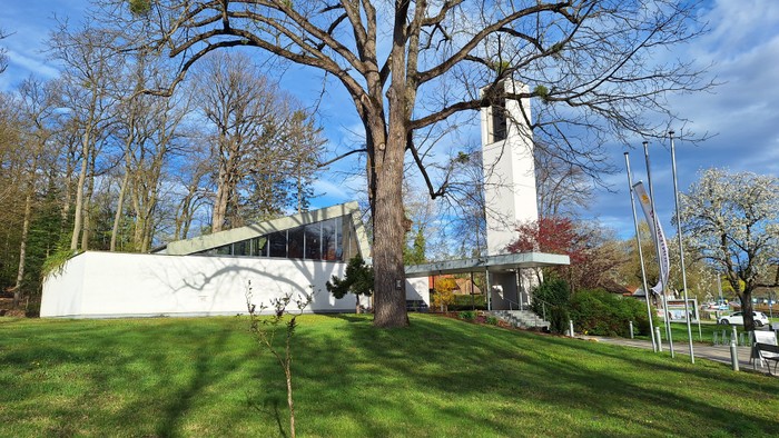 Ein weißes Gebäude mit spitzem Turm, umgeben von Bäumen und grünem Gras unter blauem Himmel