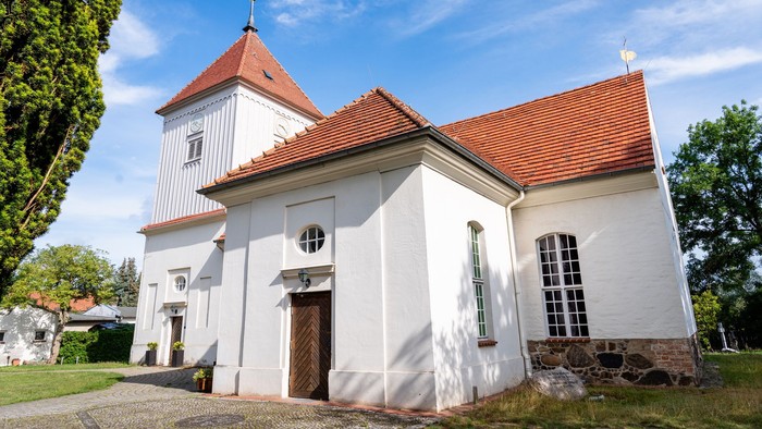 Eine charmante weiße Kirche mit rotem Ziegeldach und Glockenturm, eingebettet in eine ruhige, grüne Landschaft.