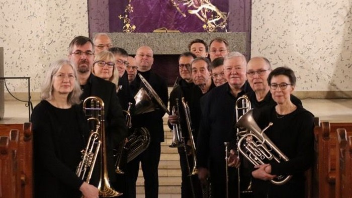 Eine Gruppe von Musikerinnen und Musikern in formeller Kleidung, die verschiedene Blechblasinstrumente halten, posiert gemeinsam in einem Innenraum.