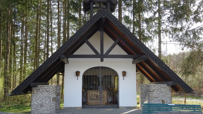 Kleines weißes Kirche in Wald mit schwarzem Dach und Holzbänken davor