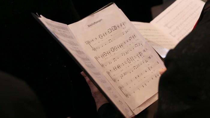 Eine ältere Dame hält ein aufgeschlagenes Liedblatt in der Hand. 