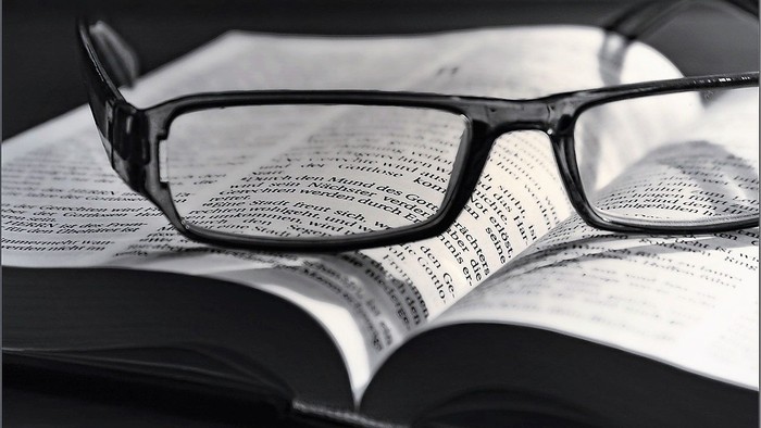 Eine Brille liegt auf einem geöffneten Buch, wobei der Text durch die Gläser sichtbar ist.