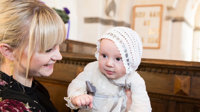 En kvinde smiler, mens hun holder en baby klædt i hvidt tøj og en strikket hue indenfor.