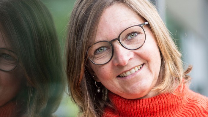 Eine fröhliche Frau mit Brille und einem orangefarbenen Rollkragenpullover lächelt in die Kamera.