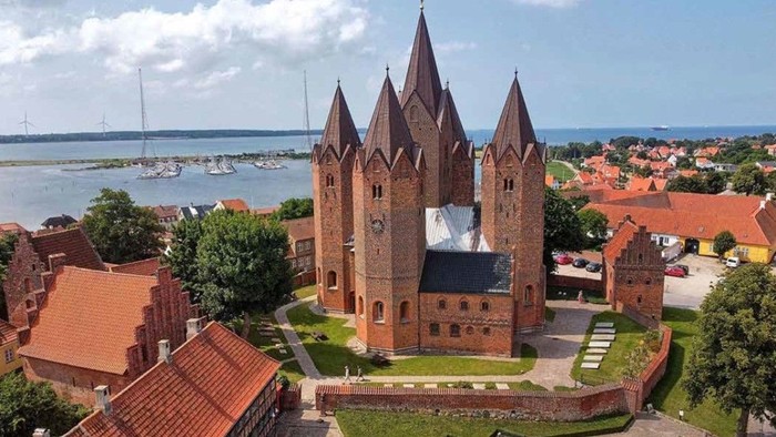 Historisk murstenskirke med spir, der kigger ud over en malerisk kystby og havn.