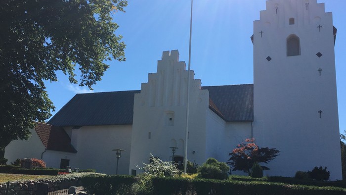Hvid kirke med tårn og klokke, omgivet af træer og mursten.