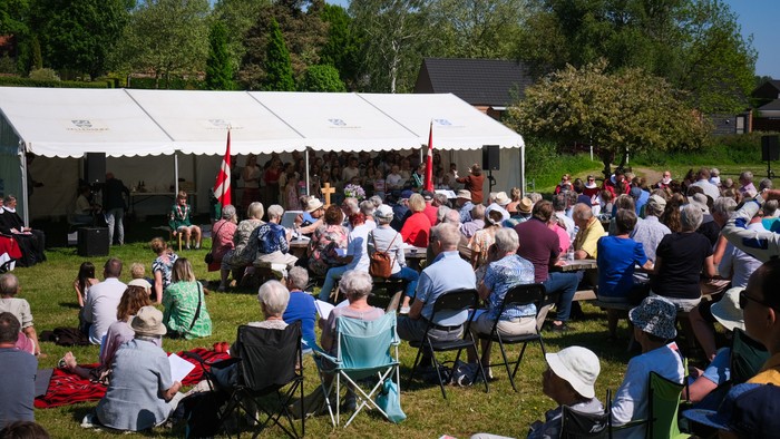 En stor gruppe af mennesker sidder på stolene foran et hvidt telt i en park.