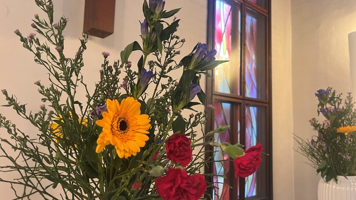 Lyse buketter med gule gerberaer, røde roser og blå blomster nær et farverigt glasmosaikvindue.