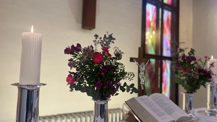 En lysende stearinlys til venstre for en vase med røde blomster og en åben bog på bordet foran et farverigt vinduesglasning.