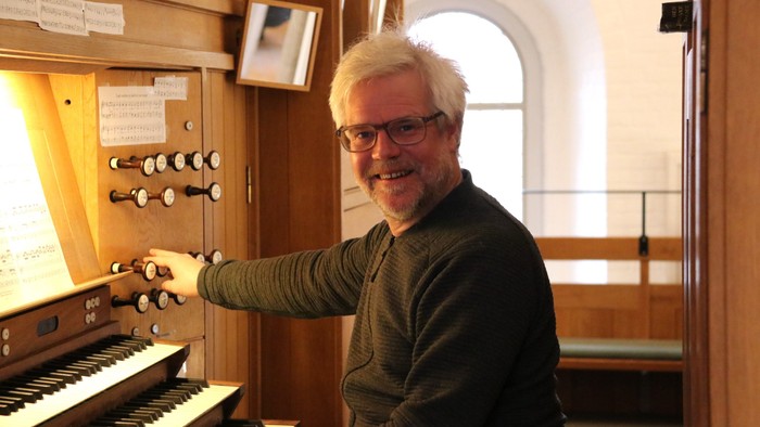 En smilende mand spiller på et orgel i en kirke med træpaneler og store vinduer i baggrunden.