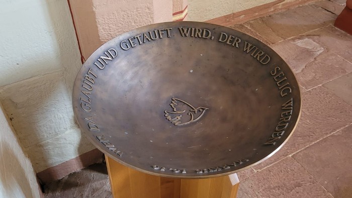 Bronzener Taufstein mit eingraviertem deutschen Text und einem Taubensymbol auf einem Holzständer.